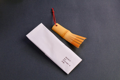 short rectangular bamboo brush with thin bristles red silk loop beside white rectangular paper wrapper with black kanji print on dark grey backdrop