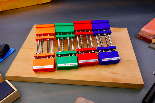four marie whetstone holders orange green red and blue sit angled atop yellowish hinoki wood cutting board surrounded by knife accessories on deep grey surface