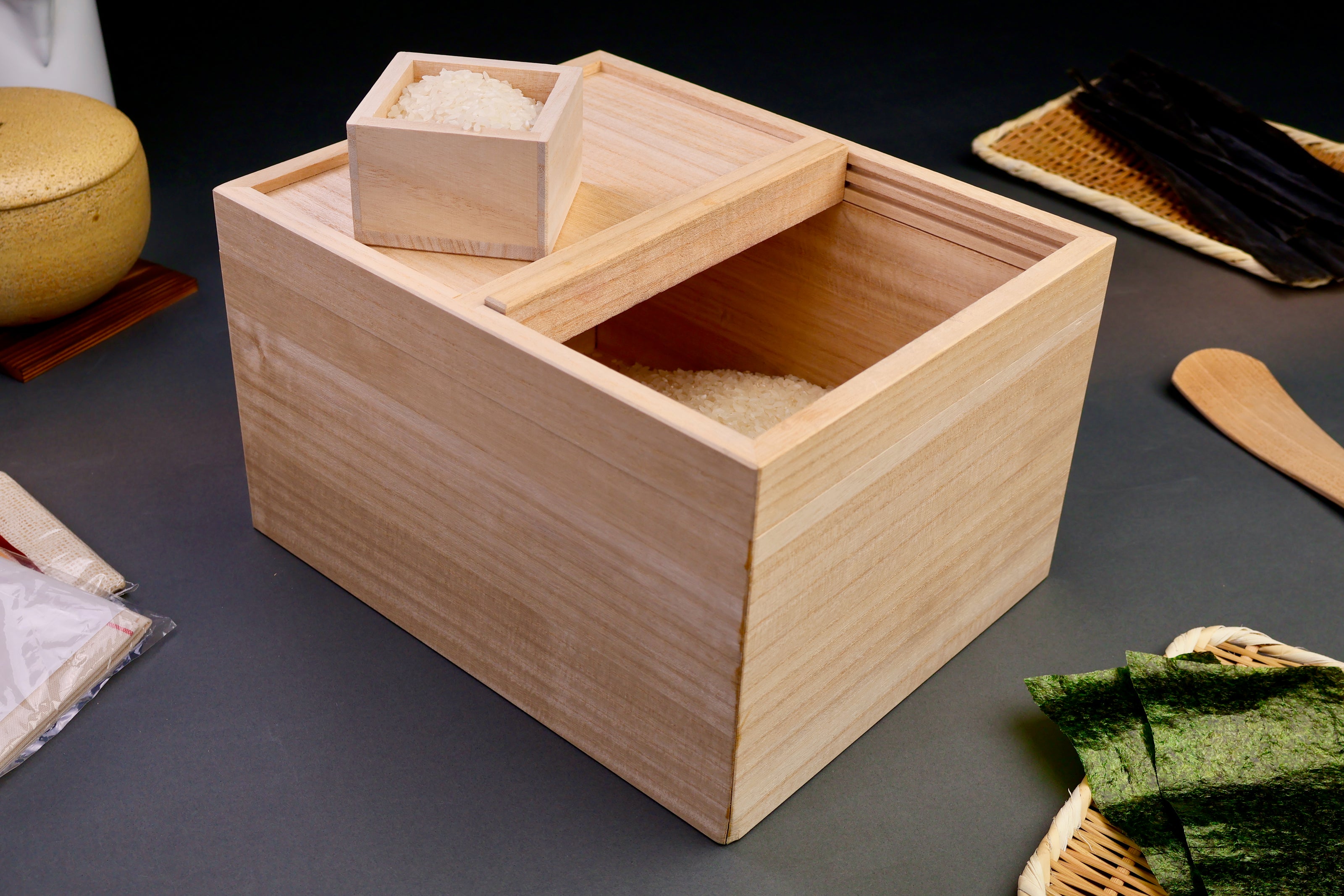 tall rectangular paulownia wood rice box sliding door on top removable with rice scoop surrounded by japanese kitchenware dark grey background