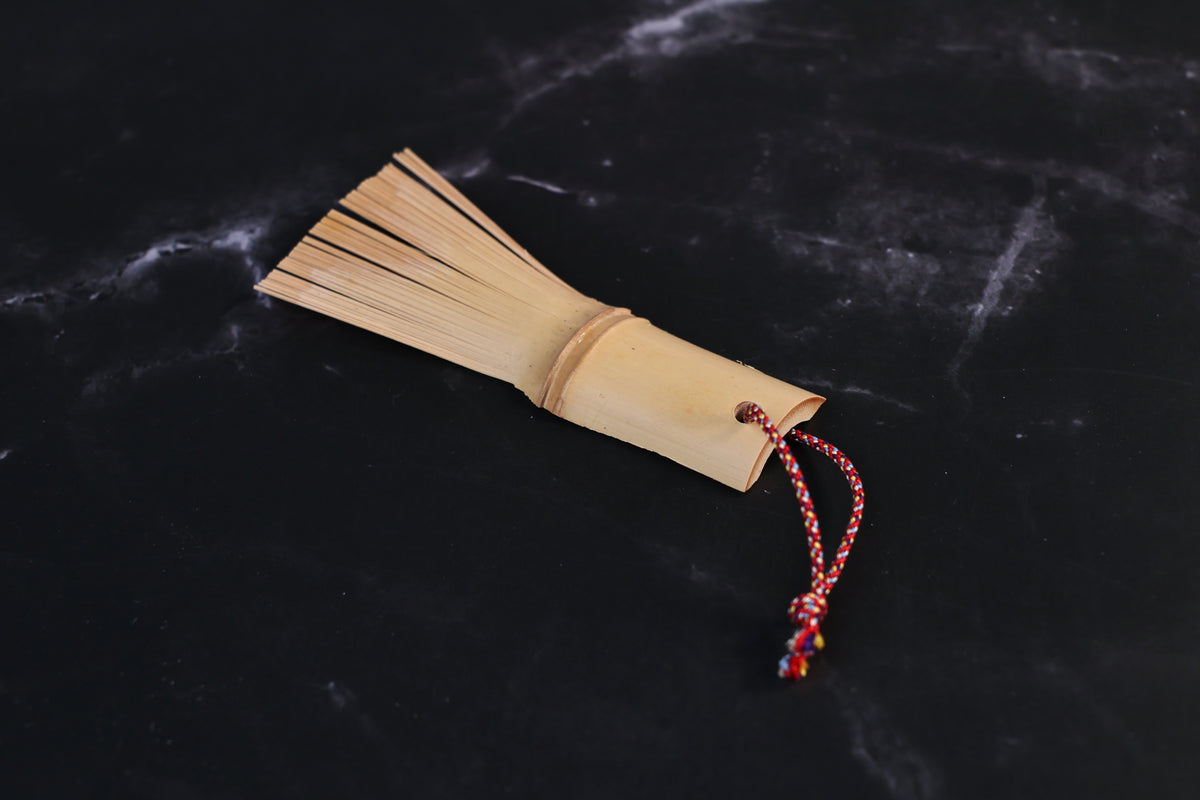 Yakumiyose bamboo brush with bristle fringe fanned out and red braided cord loop, laid on a dark surface