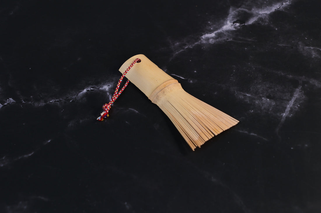 Yakumiyose bamboo brush with split bristle fringe and a red braided cord loop, shown from above on a dark surface