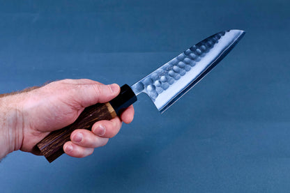 Hand holding Yoshikane SKD Tsuchime Santoku 165mm showing full blade and burnt chestnut handle grip