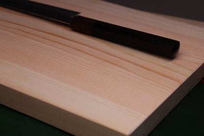 close up shot of hinoki wood grain patterns on cutting board with a chef knife on top that is dark in contrast to light pinkish colors of the wood and the very dark background